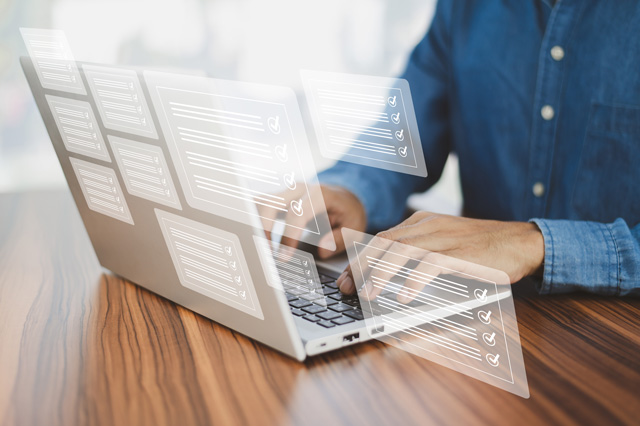 Ein Mann im Jeanshemd sitzt an einem Tisch auf dem ein Laptop steht. Der Mann tippt in den Laptop. Das Symbolbild steht für die Leistungsbestandteile der digitalen Baugenehmigung.