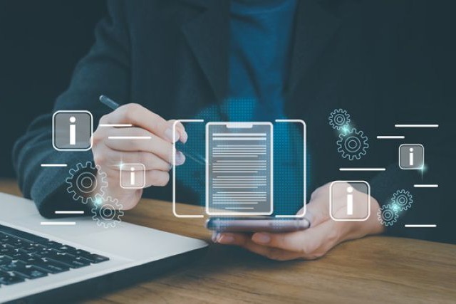 Das Symbolbild zeigt eine Person vor einem Laptop mit einem Smartphone in der Hand. Dort trägt die Person Daten ein. Das Bild soll zeigen, wie viele Menschen mit der digitalen Baugenehmigung zusammenarbeiten können.