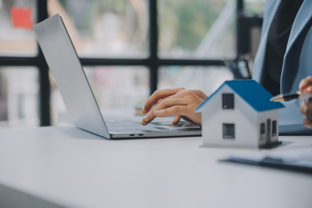 Ein Laptop steht neben einem Modellhaus auf einem Schreibtisch. Eine Person im Anzug gibt etwas in den Laptop ein. Das Bild soll eine Behörde im Umgang mit der digitalen Baugenehmigung darstellen.